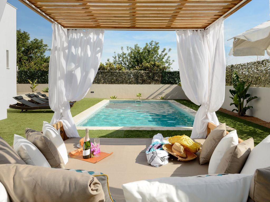 A shaded outdoor seating area is set near a private pool, with soft cushions and a decorative throw. Sun loungers are positioned alongside the pool, surrounded by greenery. Light filters through flowing white curtains, creating a calm and inviting space for relaxation.
