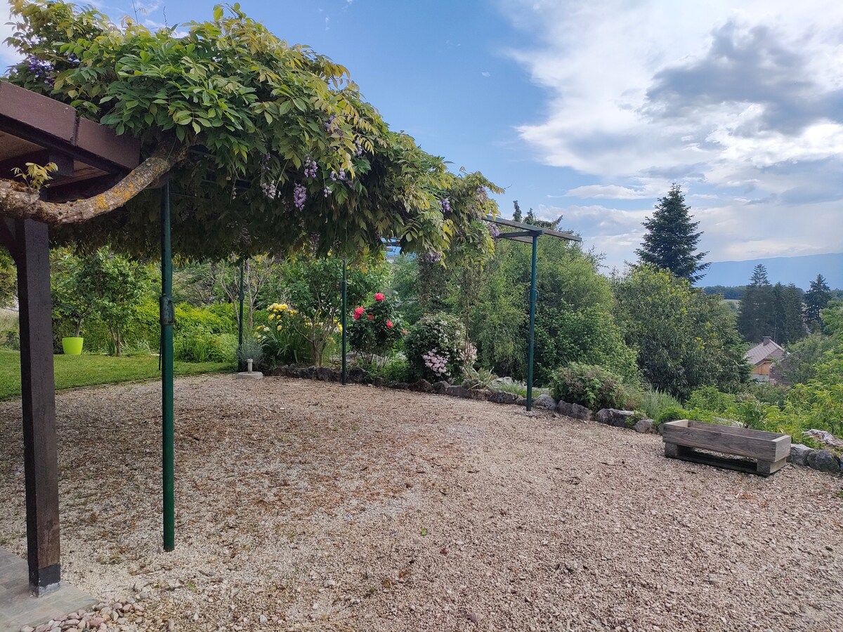 A serene outdoor space features a covered area supported by a trellis, adorned with climbing plants. Lush greenery surrounds the gravel ground, with colorful flowers adding natural beauty. In the distance, trees and hills create a calming backdrop under a partly cloudy sky.