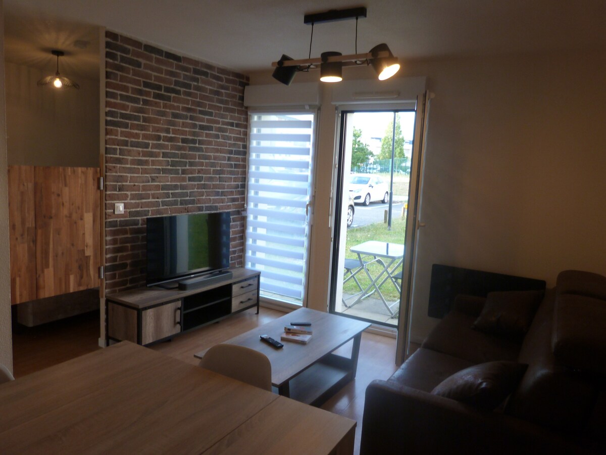 The living area features a brown leather sofa, a coffee table, and a TV on a console. A brick accent wall adds texture to the space. Natural light enters through a glass door and a window dressed with horizontal blinds, providing views of the outside.