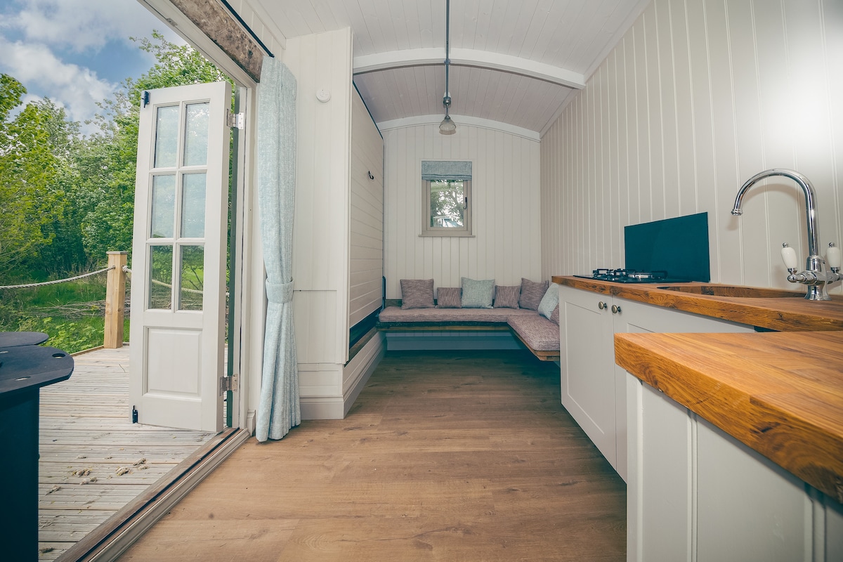 An open-plan living space features light wood flooring and white panelled walls. A corner sofa with soft cushions is positioned beside a window. A small kitchen area includes a wooden countertop, gas hob, and a sink, with doors opening onto a private deck.