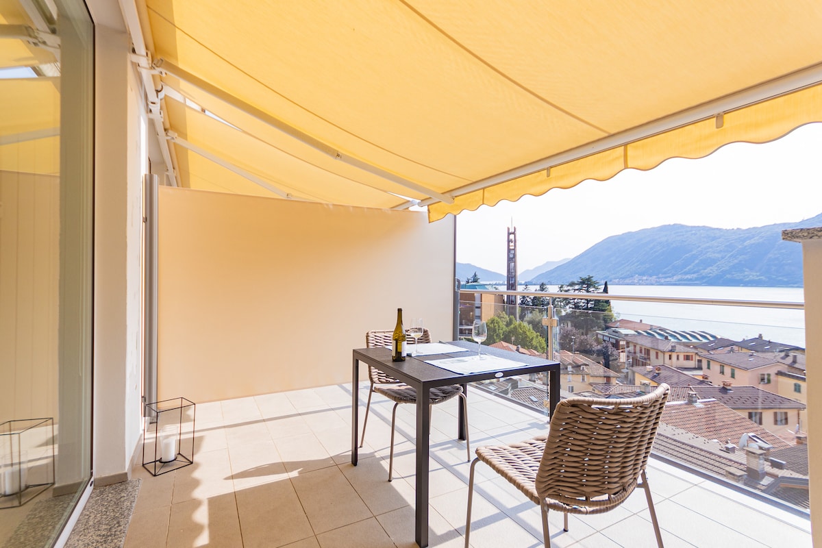 A spacious terrace features a dining table with chairs, set against a backdrop of a stunning lake view. A soft yellow awning provides shade, while the surrounding landscape includes distant hills and charming buildings. The flooring is light-colored tiles, enhancing the open atmosphere.