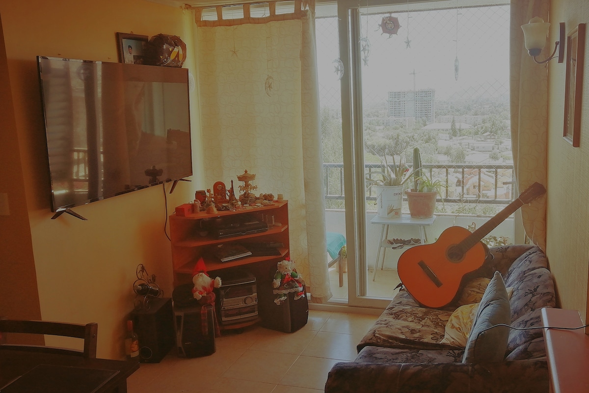 A cozy living area is featured, complete with a dark sofa and a large flat-screen TV mounted on the wall. A guitar rests beside the sofa, and a glass door leads to a balcony with a view of the city. Natural light fills the space.