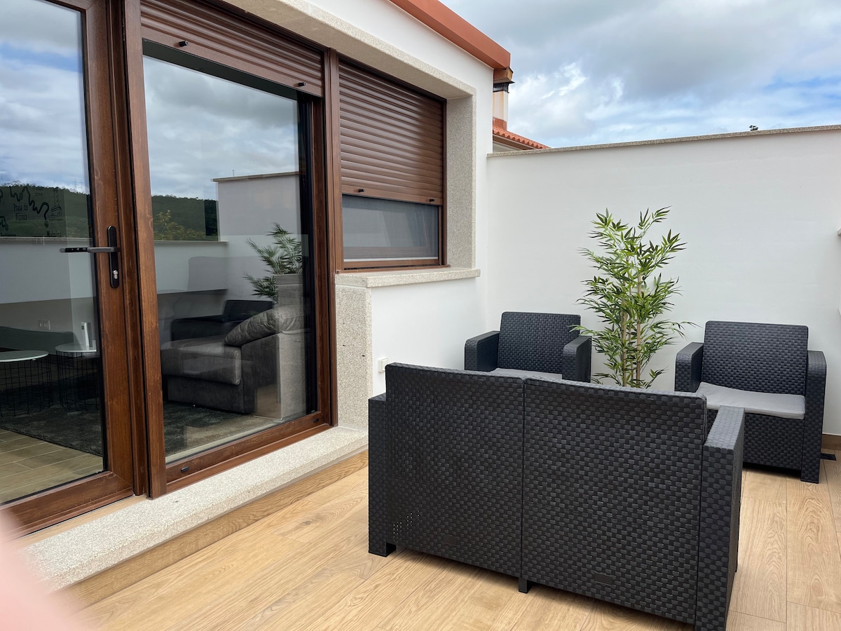 A terrace area features a comfortable seating arrangement with four dark wicker chairs surrounding a low table. Light-colored wooden flooring enhances the space, while large windows provide views of the outside. A small potted plant adds a touch of greenery to the setting.