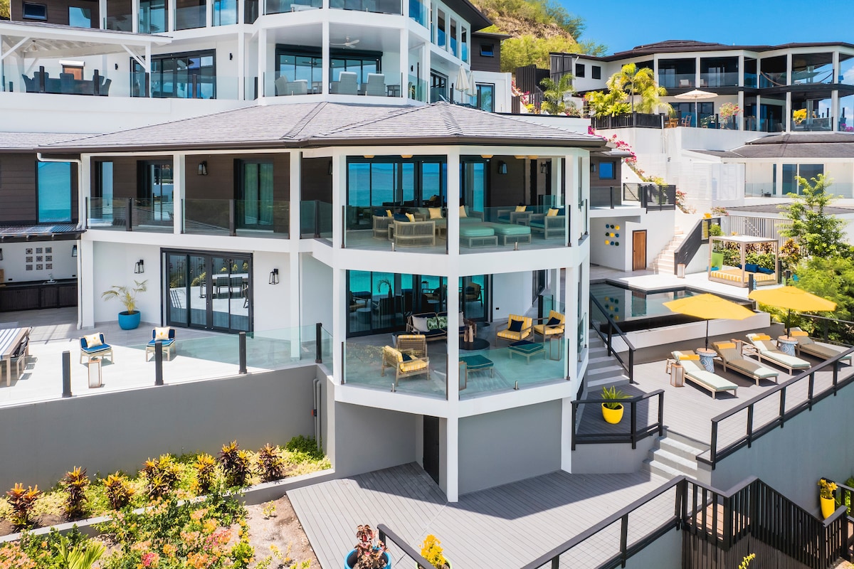 A modern multi-level home features large glass windows allowing ample natural light. Outdoor seating areas are arranged on spacious decks, complemented by yellow umbrellas. The lush landscape includes vibrant plants, adding color to the surroundings, while a staircase leads between the various levels of the property.
