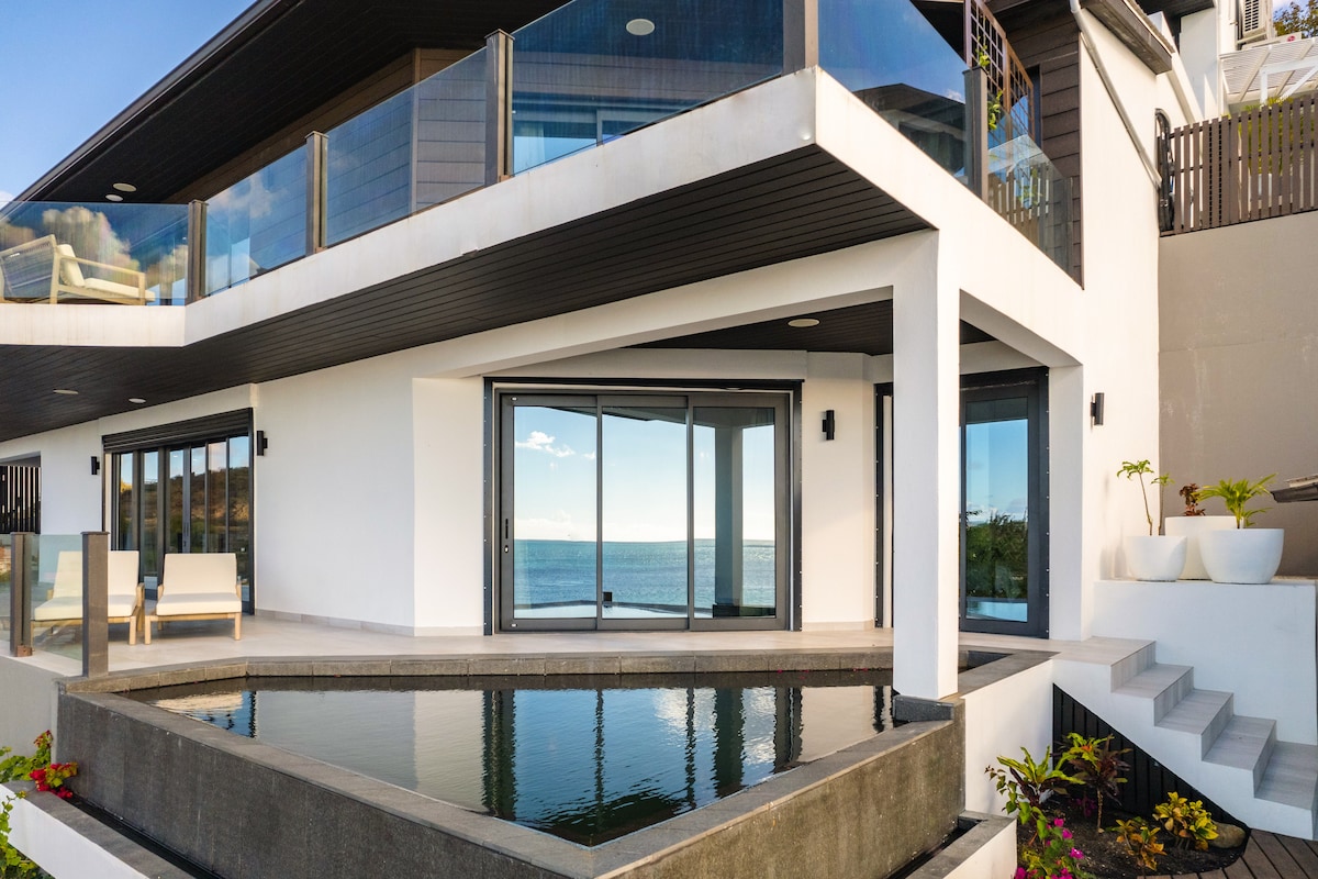 The exterior of the oceanfront home features a modern design with expansive glass doors opening to a terrace. An infinity pool reflects the sky, while comfortable seating is available nearby. Ocean views extend beyond the property, enhancing the serene environment.
