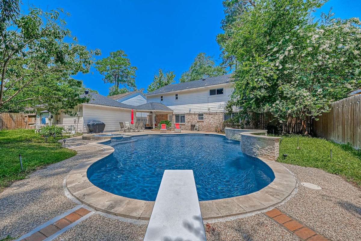A serene backyard reveals a spacious swimming pool surrounded by landscaped greenery. A concrete diving board extends into the clear water, while lounge chairs provide places for relaxation. The home features a neutral exterior blending harmoniously with the natural setting, ensuring privacy and tranquility.