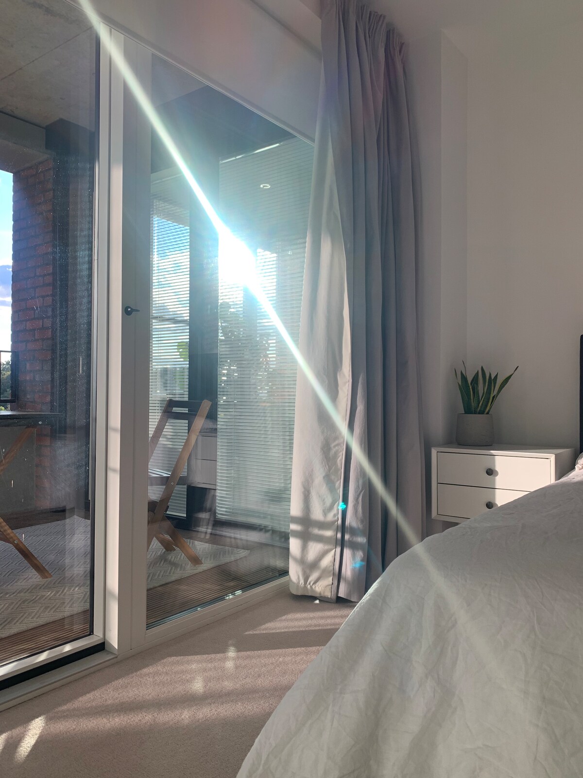 A well-lit bedroom features large glass doors that open to a private balcony, allowing sunlight to stream in. A small plant and side table are visible, while soft curtains frame the entrance. A cozy bed with a light comforter occupies the space.