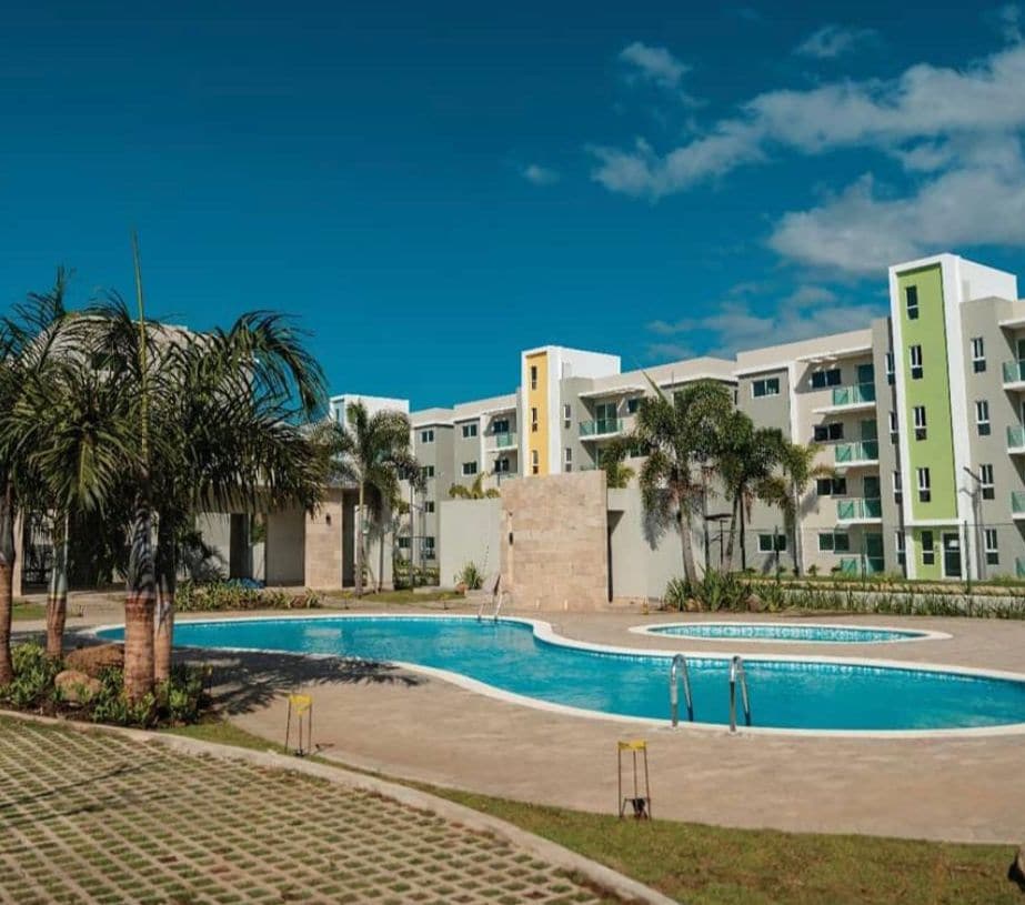 A tranquil outdoor area features a pool surrounded by lush greenery and palm trees. The building, distinguished by colorful accents, stands in the background, with clear blue skies enhancing the serene atmosphere.
