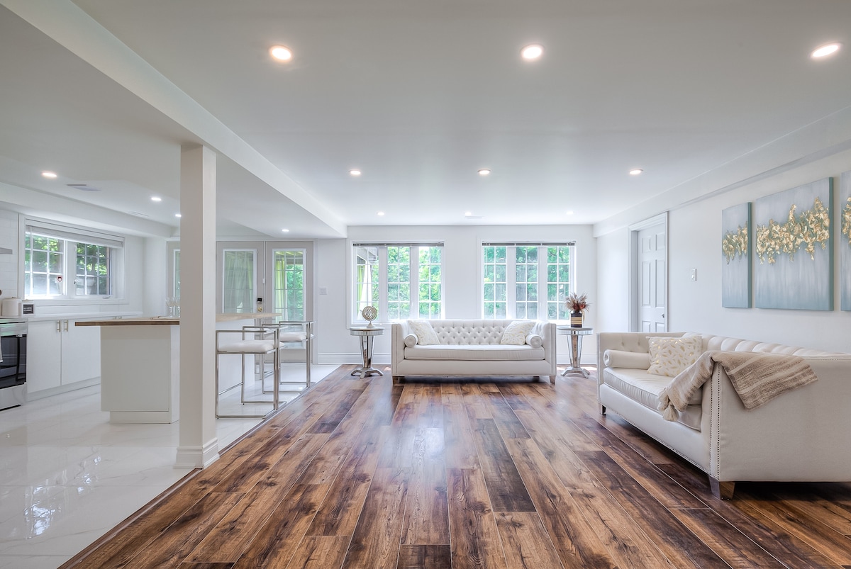 An open concept living area is showcased, featuring light-colored furniture and a spacious design. Large windows allow natural light to illuminate the room, revealing a polished wood floor. A neutral-toned artwork hangs on the wall, complementing a modern, bright kitchen visible in the background.
