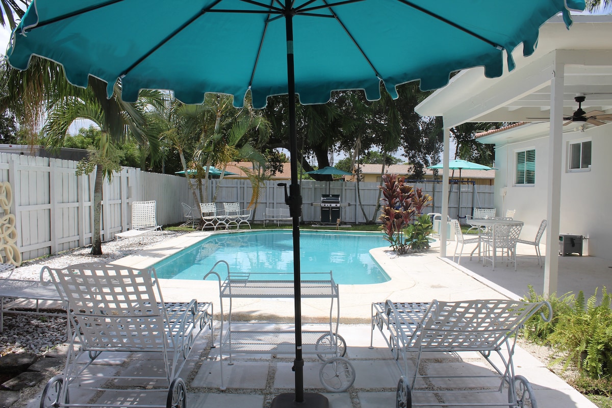 The pool area features a bright turquoise umbrella providing shade over two white lounge chairs. The pool glistens under sunlight, surrounded by landscaped greenery and a paved deck. A barbecue grill and outdoor seating are visible in the background, creating a relaxed outdoor space.
