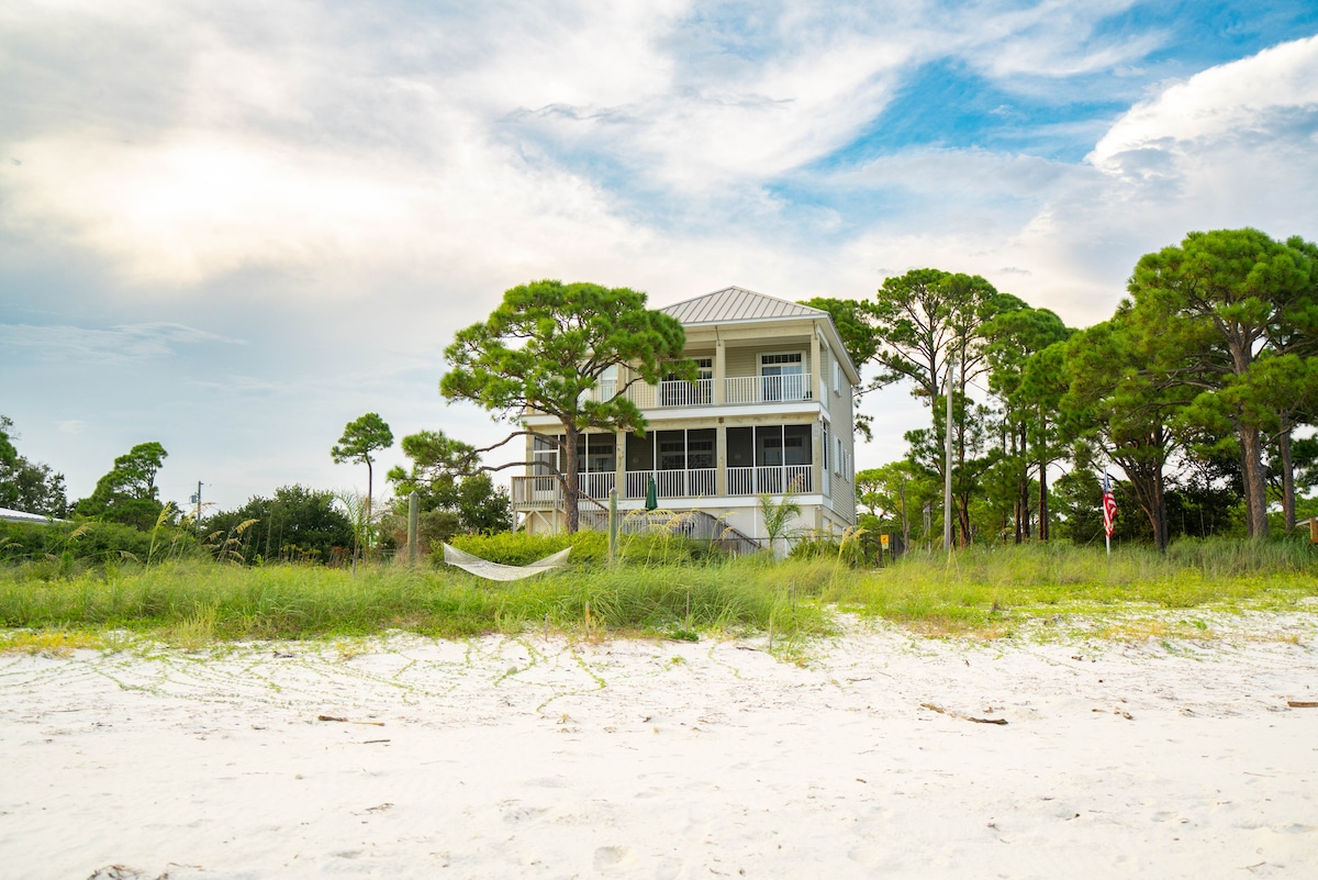 A beachfront home is set against a backdrop of green trees and cloudy blue skies. The two-story structure features a screened-in porch on the second level, surrounded by sandy grass that leads to the beach. Hammocks are visible among the trees.