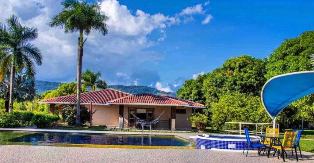 A serene outdoor area features a rectangular pool surrounded by palm trees and lush greenery. A shaded seating area with yellow chairs is visible near the pool, complemented by a hot tub and a hammock strung between two posts, all under a clear blue sky.