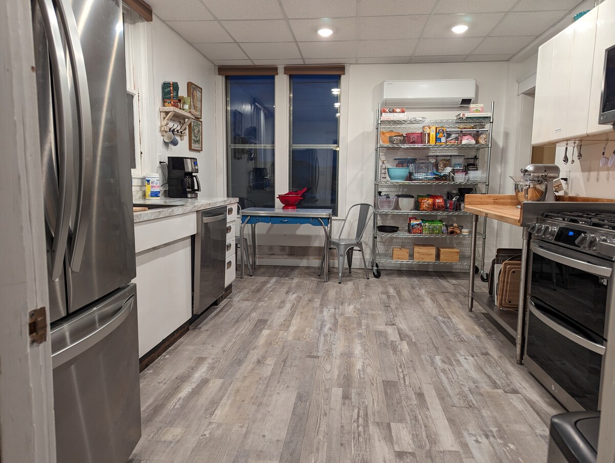 A modern kitchen is presented with stainless steel appliances, including a refrigerator and stove. A light wood counter creates a workspace, while a metal-legged table with chairs accommodates dining. Shelving displays various food items and kitchen essentials, with windows allowing natural light into the space.
