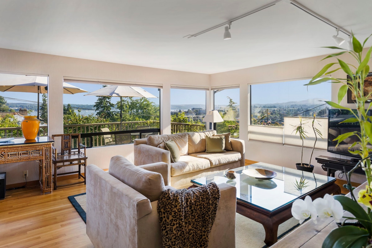 A spacious living room is showcased, featuring large windows offering expansive views of the water and mountains. Comfortable seating is provided by a beige sofa and armchair, with a coffee table featuring a decorative arrangement. Natural light brightens the inviting space, enhanced by indoor plants.