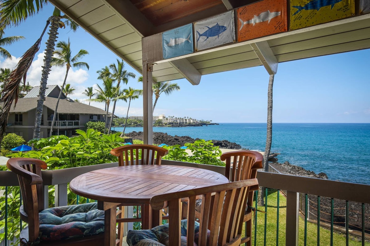 A wooden dining set occupies a private balcony, offering views of the ocean and surrounding greenery. The railing frames the picturesque coastline, with palm trees swaying gently in the breeze. Colorful artwork is displayed above, adding a vibrant touch to the serene outdoor space.