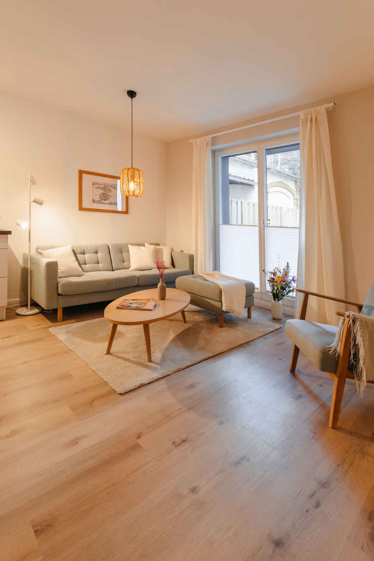 A living area features a comfortable sofa, a side chair, and a coffee table with magazines atop. Natural light enters through the glass doors, which open to a small balcony. A light color palette enhances the airy feel, while a lamp provides additional lighting.