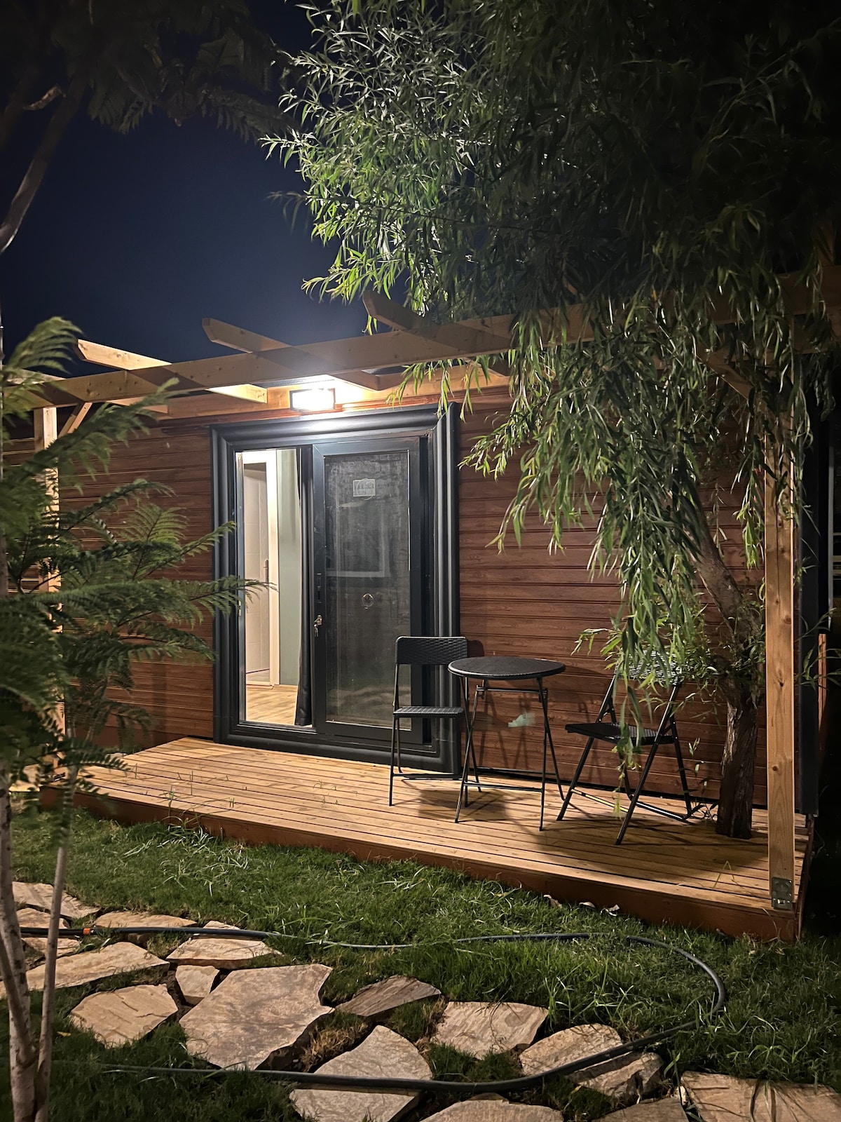 A wooden structure stands in a garden, featuring a small terrace with two black chairs and a table. Soft lighting illuminates the entrance, creating a cozy nocturnal ambiance. Surrounding greenery adds a touch of nature to the setting.