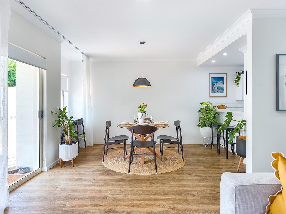 An open-plan dining area features a round table set for six, surrounded by black chairs. Large windows allow natural light to enhance the space, complemented by indoor plants. A warm area rug lies beneath the table, creating a cozy ambiance. The adjacent kitchen is visible in the background.