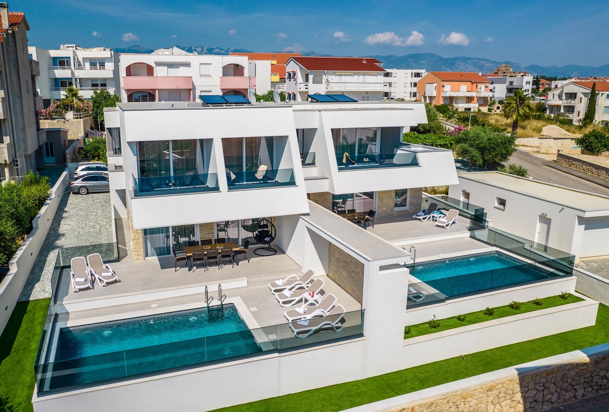 The villa features a modern architectural design with two distinct sections. Each section contains a private pool surrounded by lounge chairs. The spacious terraces with glass railings provide an unobstructed view of the surrounding area, complemented by neatly manicured greenery.