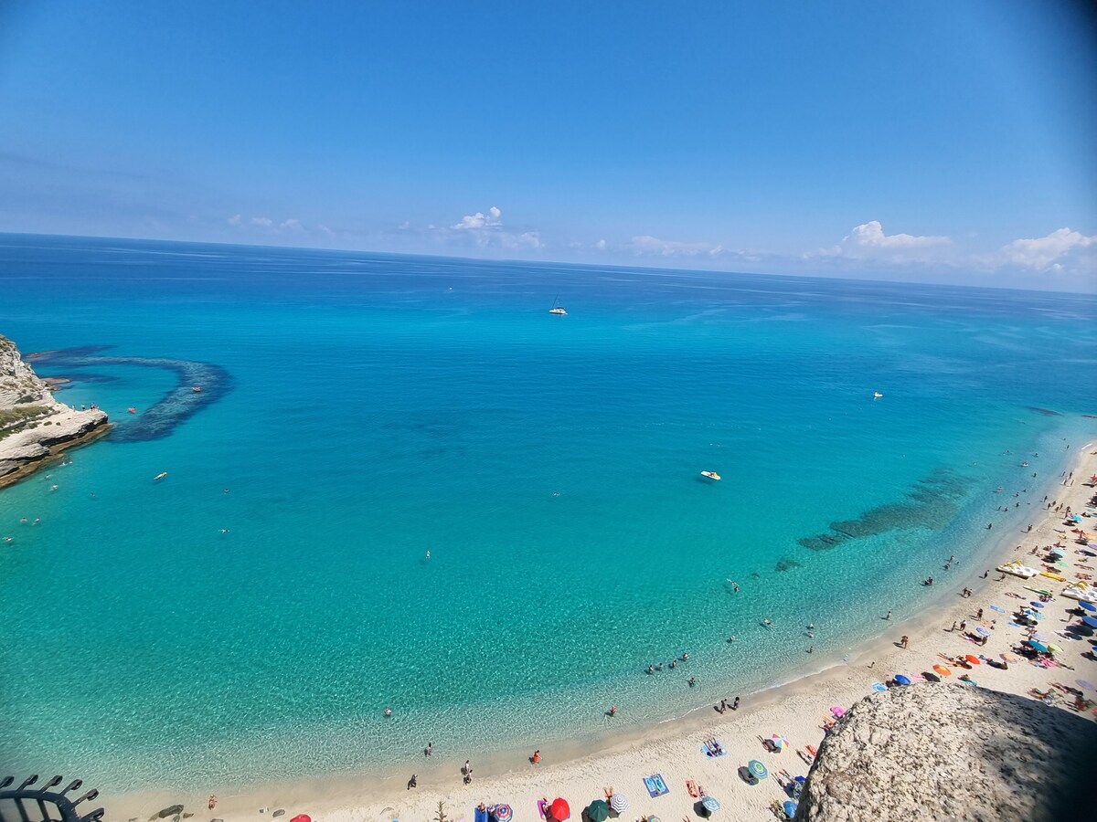 A wide view of a sandy beach with colorful umbrellas scattered across the shore. Clear turquoise waters extend to the horizon, revealing gentle waves and a few boats. The sky is bright blue with wispy clouds, enhancing the overall coastal scene.