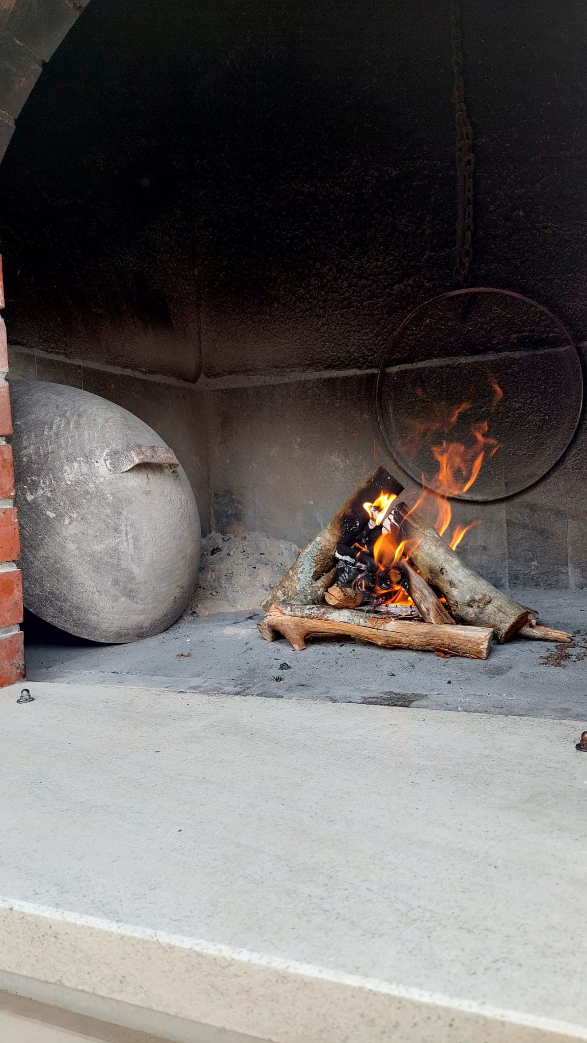 A stone barbecue grill is showcased, featuring a small fire with flames flickering among logs. A round, metallic lid rests against the side, contributing to the rustic appearance. The grill's interior is lined with dark stone, enhancing the overall cooking space.