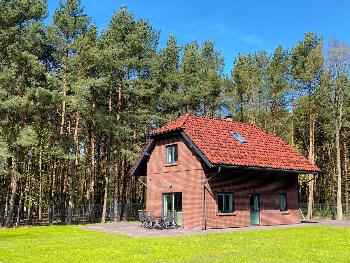 A two-story villa is situated in a lush green setting, surrounded by tall trees. The building features a red tiled roof and multiple windows. A patio area with outdoor furniture is visible in front, set against an expansive grassy lawn.