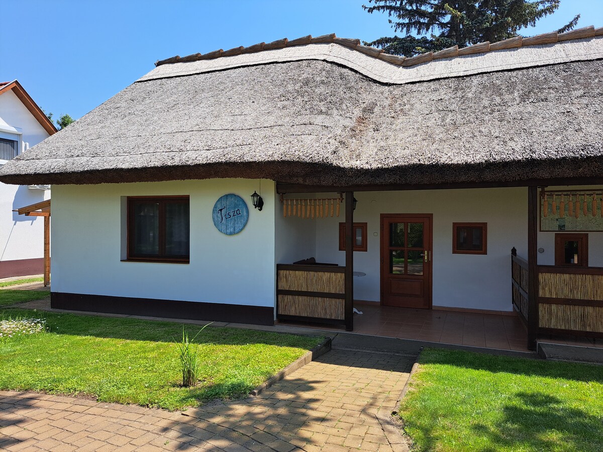A traditional thatched-roof house is set amidst a lush green lawn. The entrance features a wooden door with a decorative sign. Pathways of stone lead to the front, bordered by well-maintained grass and shrubs.
