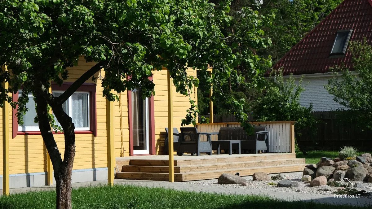 An exterior view of a bright yellow house features large windows and a welcoming entrance. A wooden terrace furnished with outdoor seating is shown, surrounded by greenery and stones. A tree partially shades the area, adding to the relaxed ambiance.
