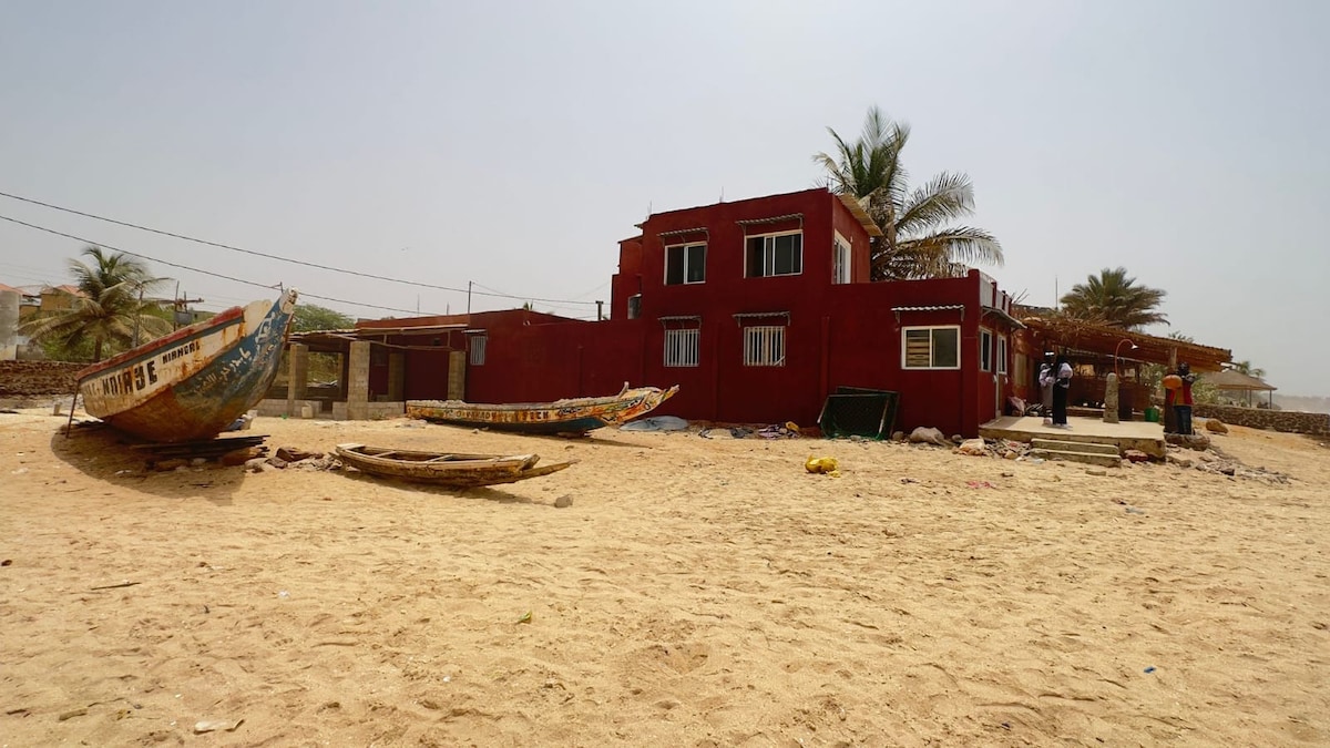 The exterior of the house is viewed against a sandy beach, featuring a red façade with multiple windows and a patio area. Two traditional fishing boats rest in the foreground. Palm trees are visible, adding to the coastal ambiance.