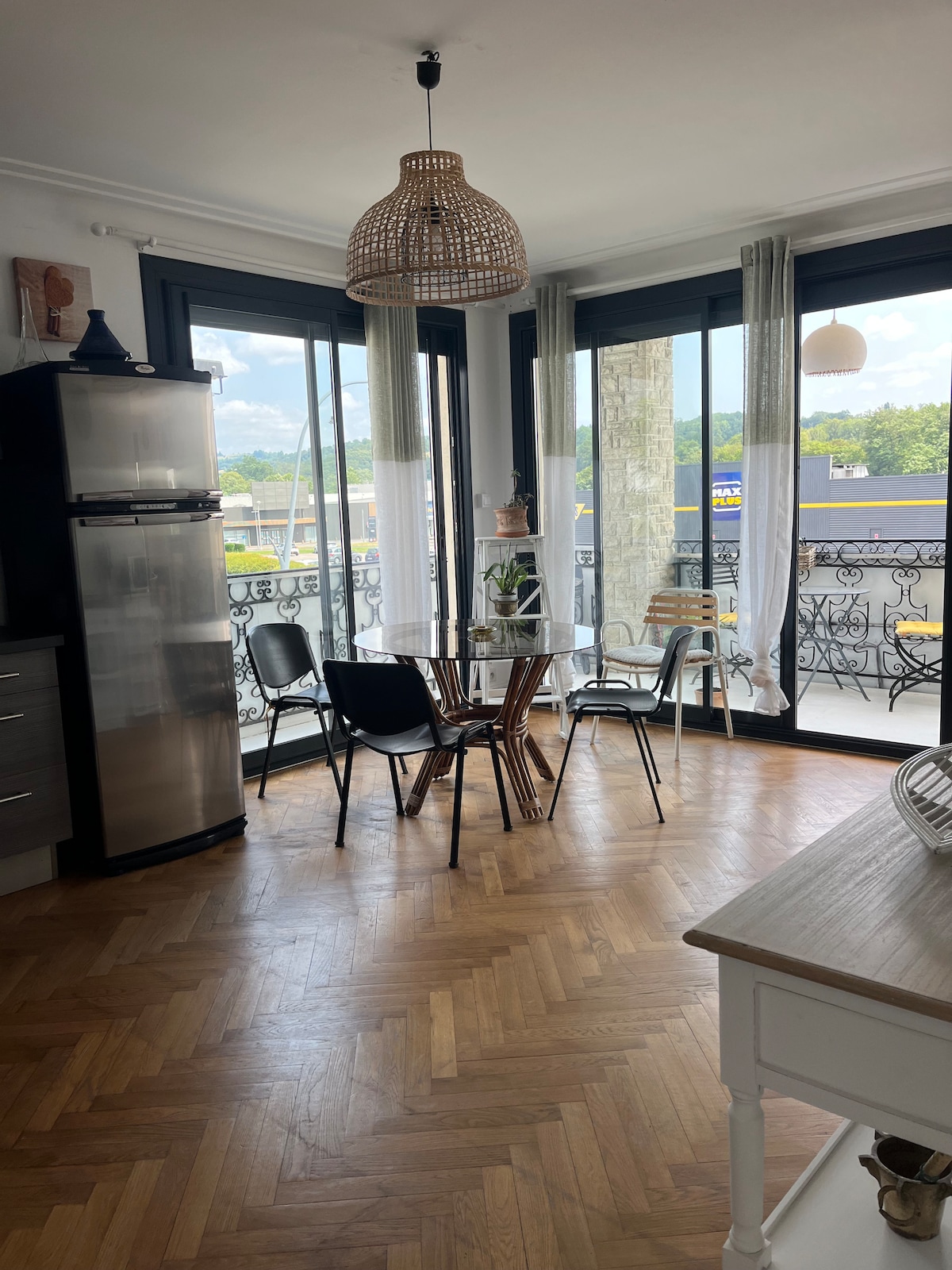 A bright dining area features a round wooden table surrounded by six black and wooden chairs. Large windows allow natural light to fill the space, offering views of the outdoors. A stylish pendant light hangs above, complementing the warm wooden flooring.