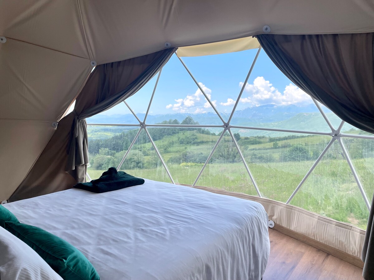 A spacious glamping dome interior features a comfortable bed, with a large front window providing views of the surrounding mountains and greenery. Natural light fills the space, while soft drapes frame the window, creating an inviting atmosphere.