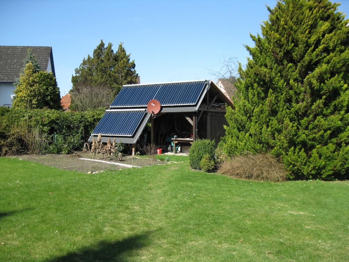 A spacious garden is visible, featuring a sunlit area with green grass. A structure covered with solar panels is nestled among shrubs and trees, with a satellite dish mounted on the roof. The scene is framed by well-maintained greenery.
