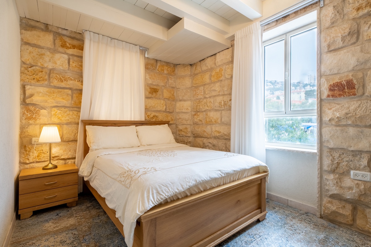 A serene bedroom features a wooden bed with a simple white bedding set. Soft light is provided by a bedside lamp next to a wooden nightstand. Textured stone walls create a cozy atmosphere, while a window allows natural light to filter in.