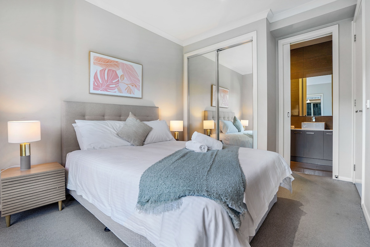 A comfortable bedroom features a spacious bed with a plush headboard. Nightstands with lamps are placed beside the bed. A large mirror reflects the en-suite bathroom, adding depth to the space. Soft textiles and subtle decor create a serene ambiance.