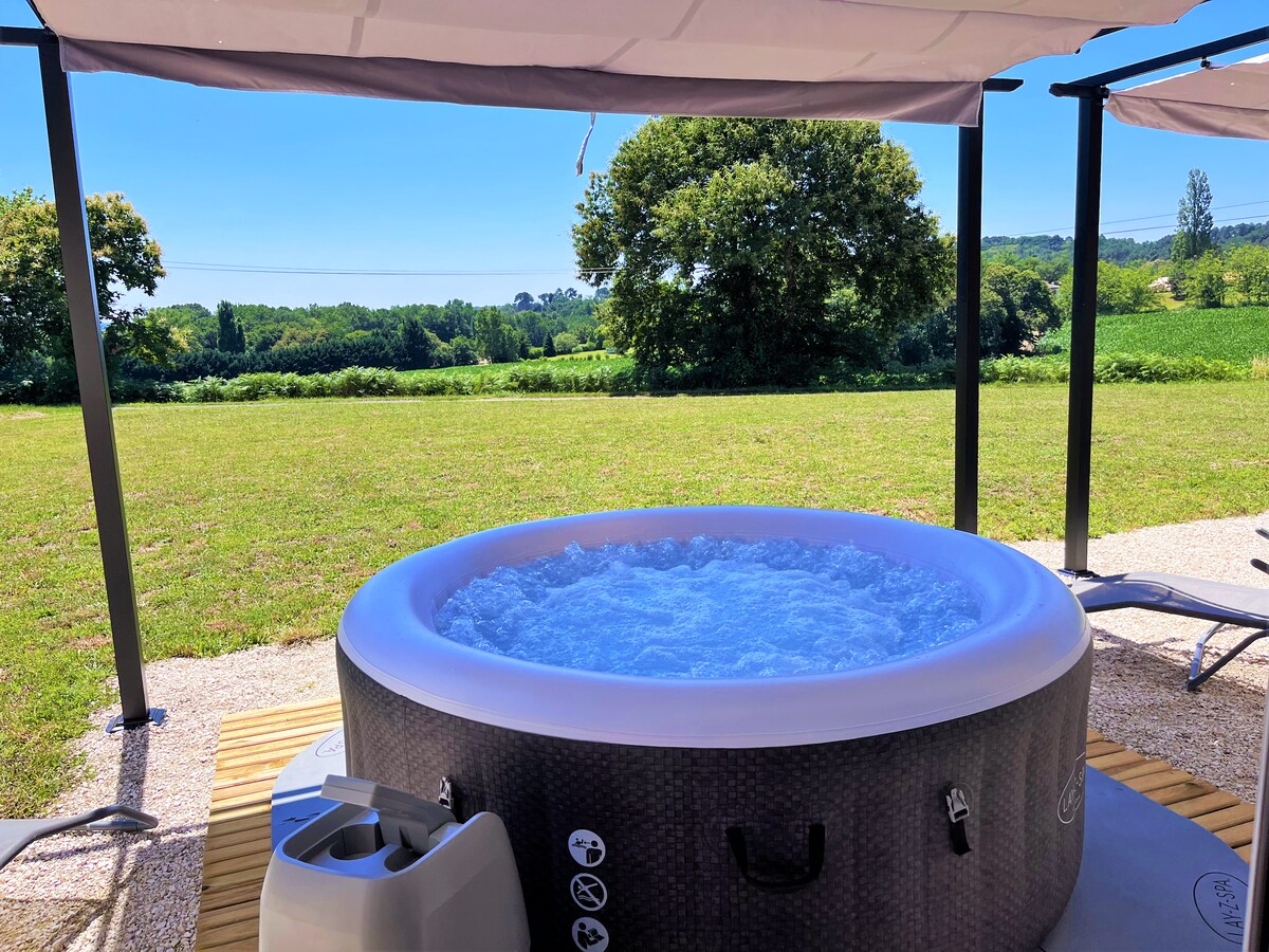 A relaxing outdoor jacuzzi is positioned under a shaded gazebo. The clear water ripples gently, inviting moments of leisure. A lush green landscape extends in the background, framed by trees and open fields, offering a sense of tranquility.