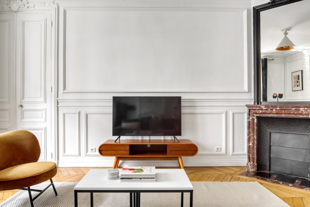 The living area features a modern television placed on a wooden console, complemented by a plush orange armchair. A minimalist coffee table sits in front, with neutral tones enhancing the space's elegance. Detailed mouldings and a fireplace add character to the room.