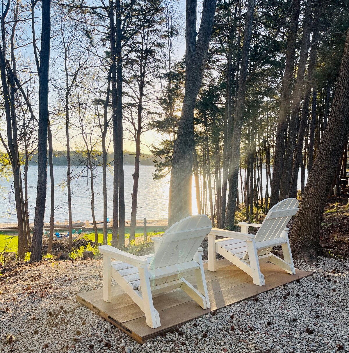 Two white Adirondack chairs are positioned on a wooden deck, facing a serene lake surrounded by trees. Soft sunlight filters through the branches, illuminating the area and reflecting off the water's surface.