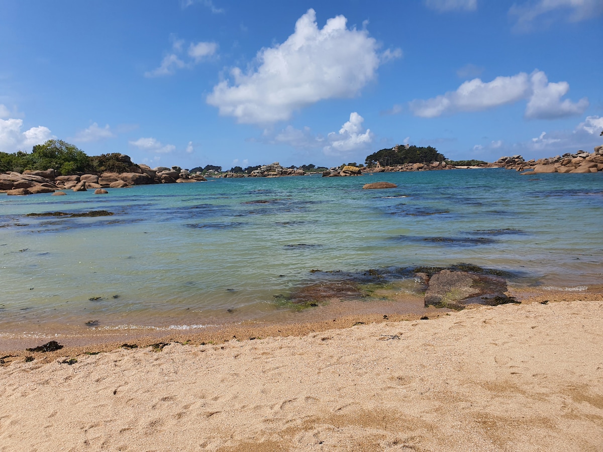 A sandy beach extends towards clear, blue waters, displaying patches of green and rock formations in the distance. Fluffy white clouds hover above, framing a serene coastal landscape that invites exploration.