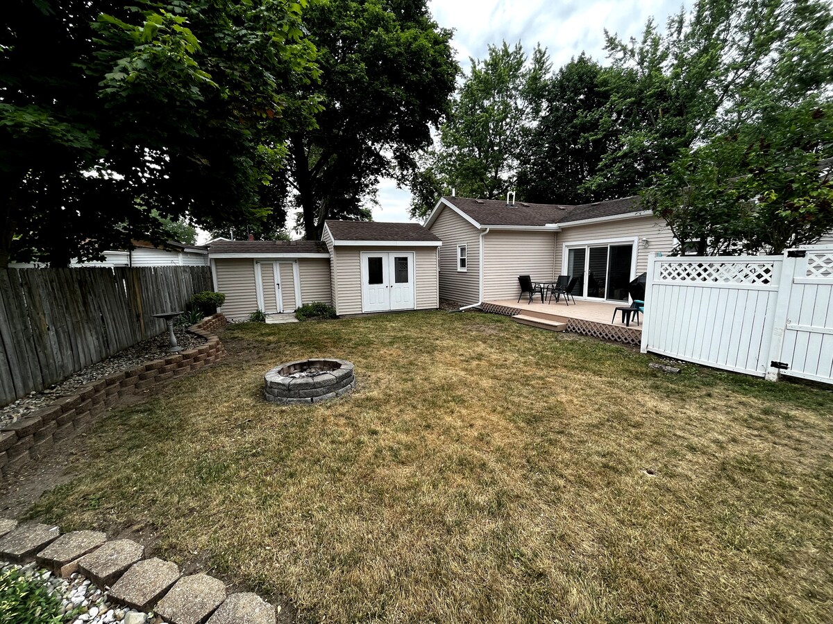 The backyard features a well-maintained lawn with a circular stone fire pit surrounded by gravel. A wooden deck is visible in the background, with multiple chairs set around a table. A storage shed is located on the left, framed by trees and a fenced perimeter.