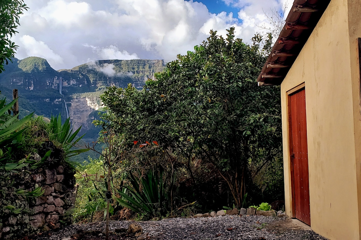 A serene outdoor scene is presented, showcasing lush greenery and surrounding mountains. The cottage’s rustic exterior is visible, framed by vibrant foliage. Clouds hover above, creating a peaceful atmosphere, while a gravel path leads to the entrance.