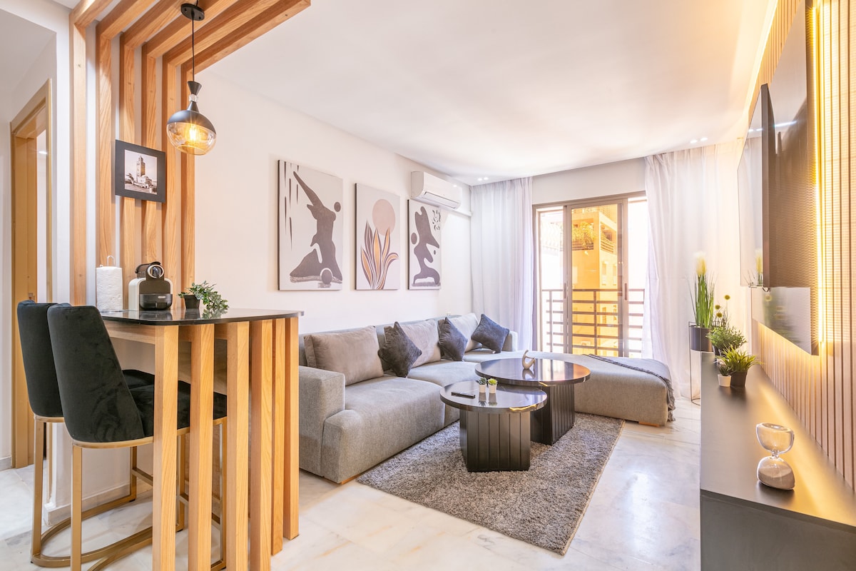 A modern living area is featured, with a spacious grey sofa and minimalist coffee table positioned on a light-colored rug. Large windows allow natural light to brighten the space, and a dining area with sleek bar stools is visible, complemented by warm wooden accents.