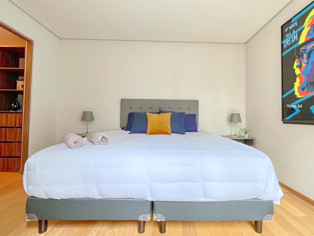 A spacious bedroom features a large bed with a neutral-colored quilt and decorative pillows in blue and yellow. Two bedside lamps are positioned on either side of the bed, and a small plant adds a touch of greenery. A piece of wall art is visible, providing a pop of color.