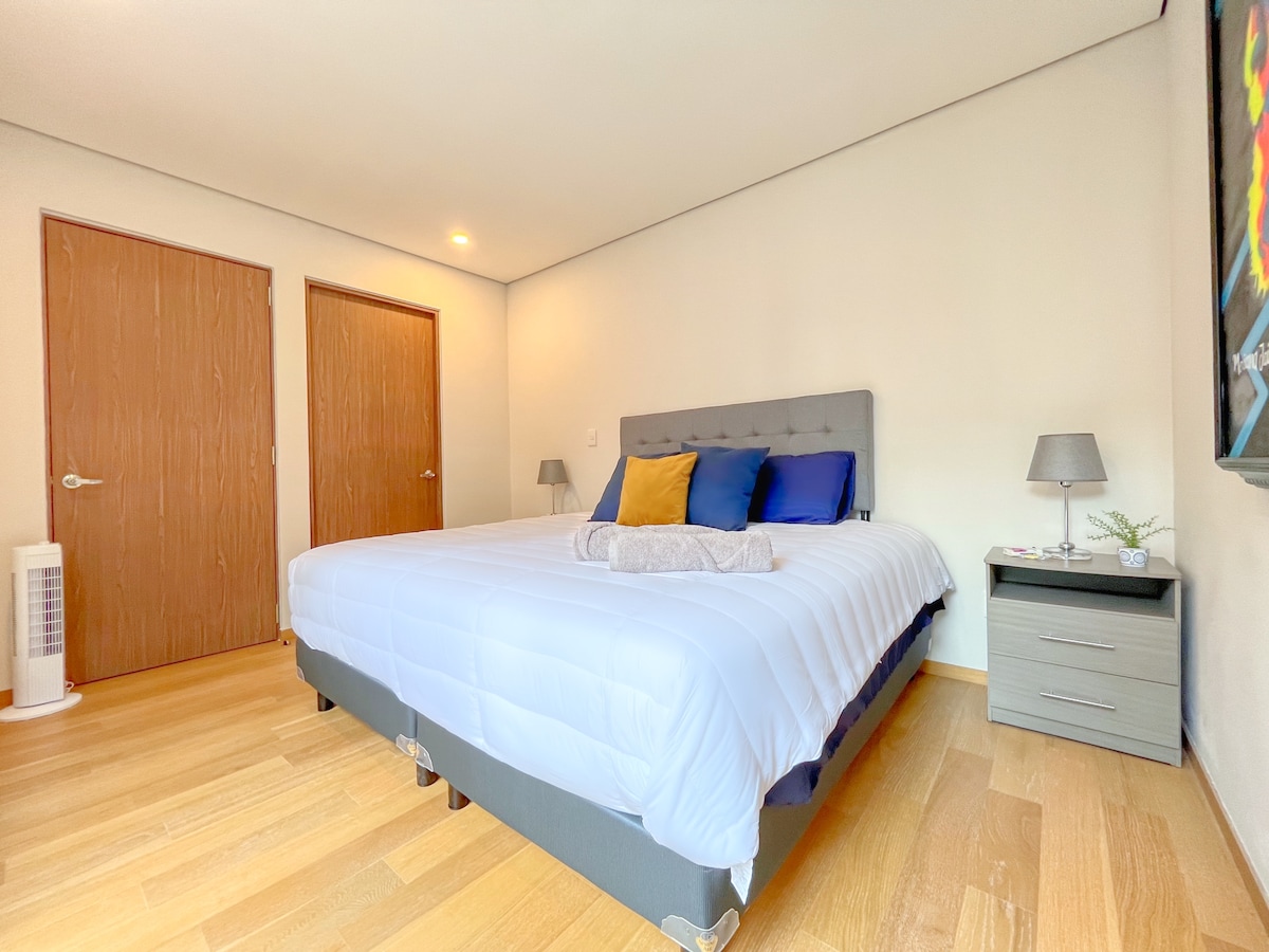 A spacious bedroom features a large bed adorned with a light-colored duvet and decorative pillows. Two wooden doors are visible, along with a bedside table that holds a small lamp. Subtle artwork adds a pop of color to the neutral walls.