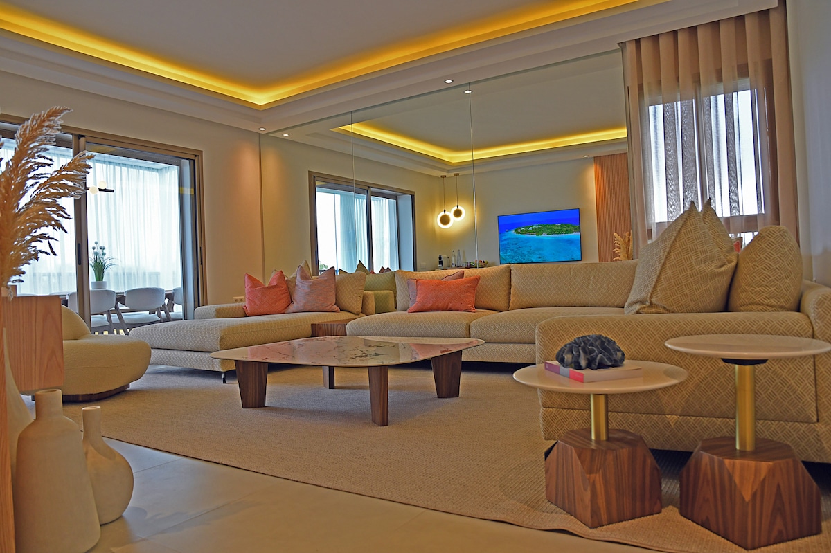 A spacious living area is filled with natural light from large windows, featuring a soft sectional sofa adorned with decorative cushions. A low, round coffee table sits atop a light rug, complemented by unique wooden side tables. A modern television is mounted on the wall.