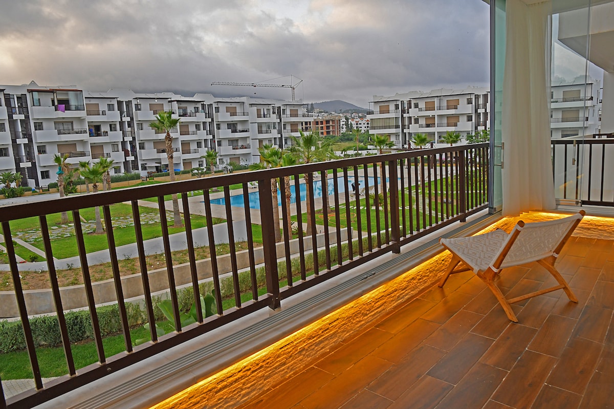 A private balcony is depicted with a comfortable chair positioned to overlook the landscaped gardens and two swimming pools below. The surrounding modern buildings contrast with the greenery, while soft lighting enhances the relaxing atmosphere of the space.