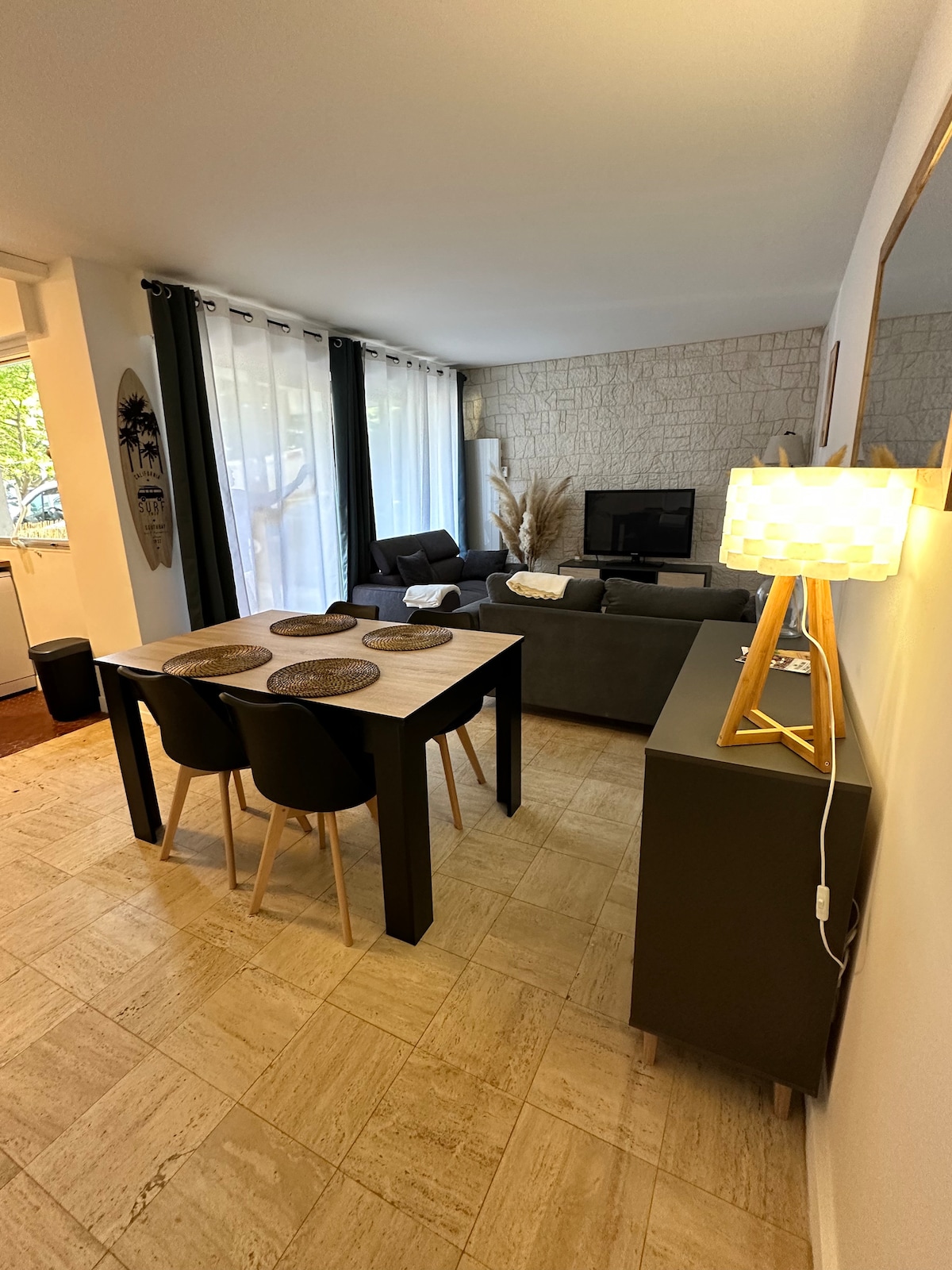 A cozy living area features a dining table with four chairs, complemented by decorative placemats. A comfortable gray sofa is positioned nearby, along with a television mounted on the wall. Soft light is provided by a standing lamp, enhancing the inviting atmosphere.