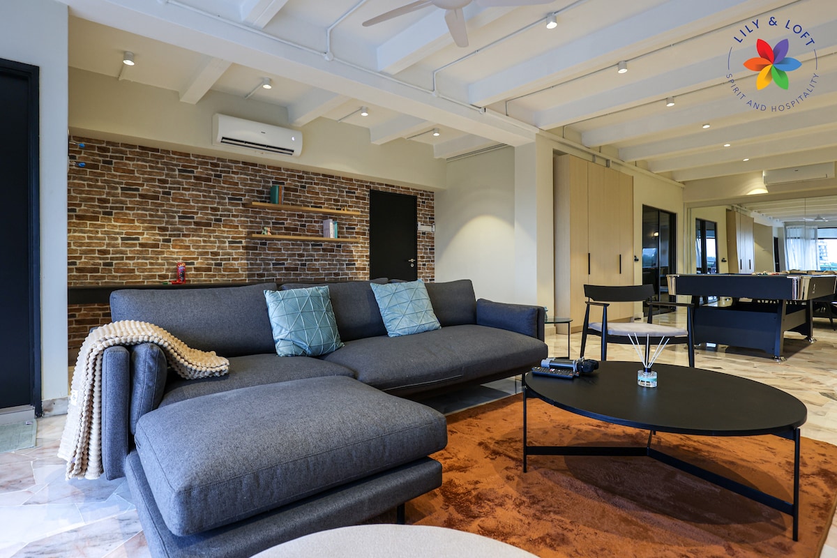 A spacious living area features a large gray sectional sofa accented with colorful cushions. A round coffee table with a dark finish sits at the center. Textured brick walls provide a modern contrast to the sleek design, while natural light fills the room through large windows.