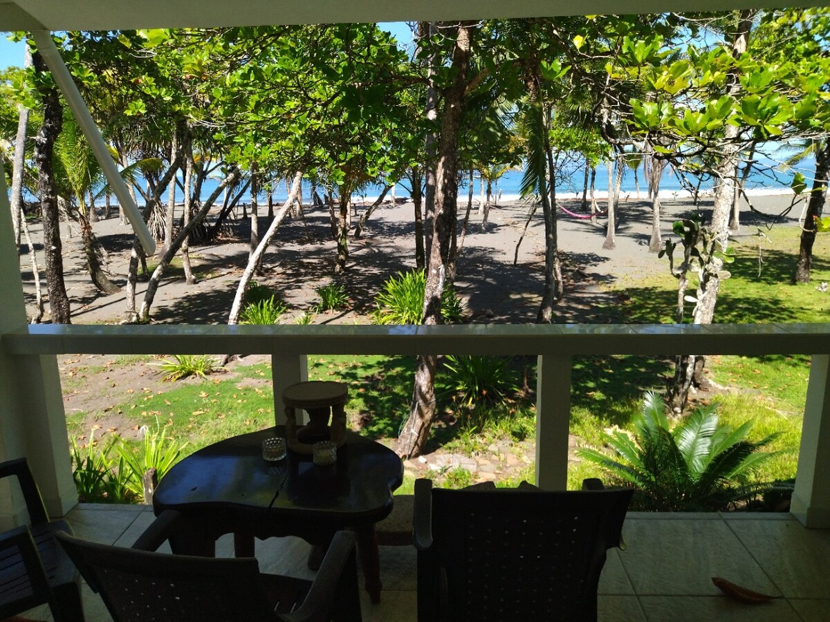 A view of a sandy beach is framed by lush greenery and palm trees. The foreground features a dark wood table and matching chairs on a covered patio, providing a shaded spot to enjoy the landscape and ocean. Sunlight filters through the leaves, enhancing the serene setting.