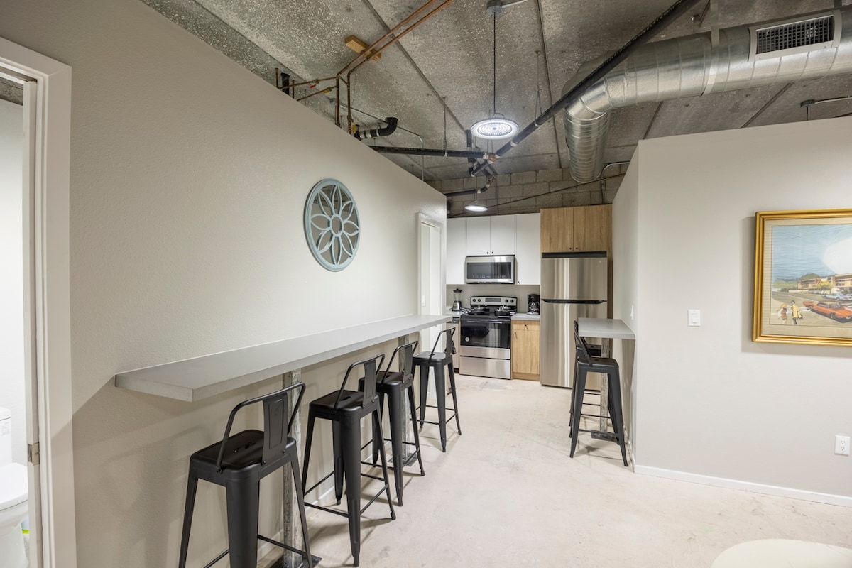 A modern kitchen space features sleek cabinetry and stainless steel appliances. A long bar with high stools creates an inviting dining area. Soft lighting illuminates the space, with a round wall decoration adding a subtle accent.