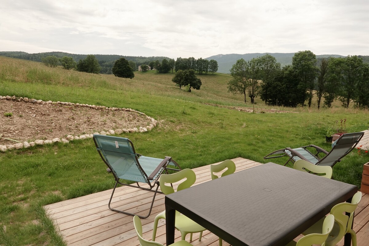 An outdoor terrace features a rectangular dining table surrounded by colorful chairs. Two lounge chairs are positioned for relaxation, facing a scenic view of rolling hills and distant trees. The open space invites an appreciation of the surrounding nature.